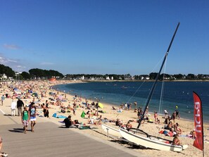 Ein voller Strand mit Menschen, die sich sonnen und schwimmen, ein Segelboot liegt im Sand, und ein klarer, blauer Himmel.