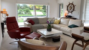 Een moderne woonkamer met een rode leren fauteuil, een witte bank en een salontafel. Op de muur hangt een scheepsschroef en er is uitzicht op groen.
