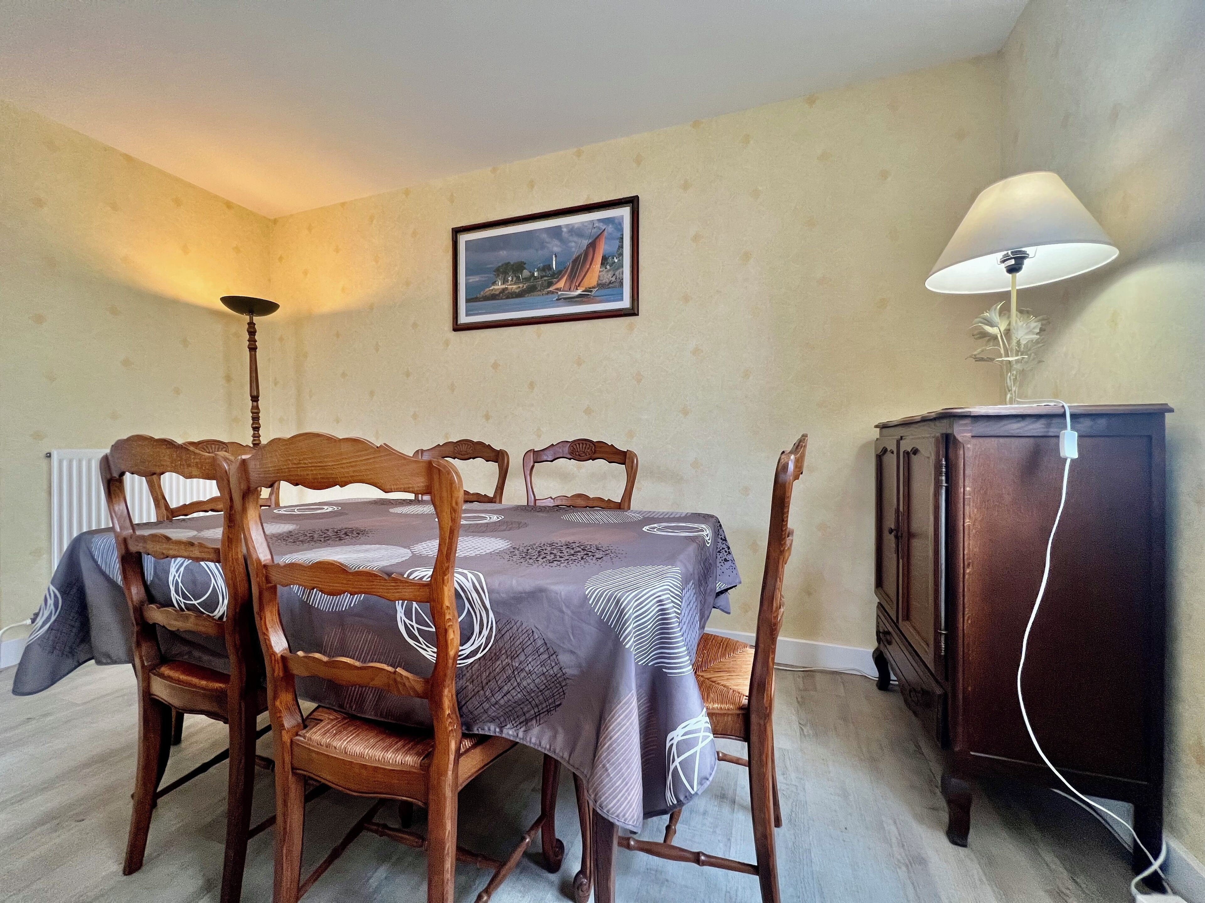 A dining room with a table set for four, wooden chairs, a framed picture on the wall, a lamp, and a cabinet with a power cord.