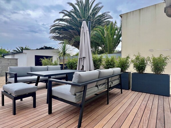 An outdoor patio with a wooden deck, grey cushioned seating, and a large umbrella.