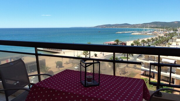 Un balcón con una mesa roja, sillas y vista al mar y la playa.