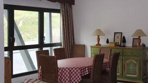 A dining area with a table set for four, wicker chairs, a green cabinet with decorative items, and a window with curtains.