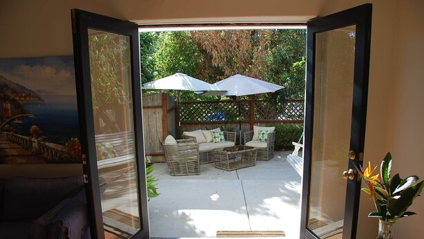An open glass door leading to an outdoor patio with wicker furniture, a white umbrella, and a view of a coastal landscape. - Hidden Villa- A Hidden Paradise with beautiful furnished garden patio (San Diego)