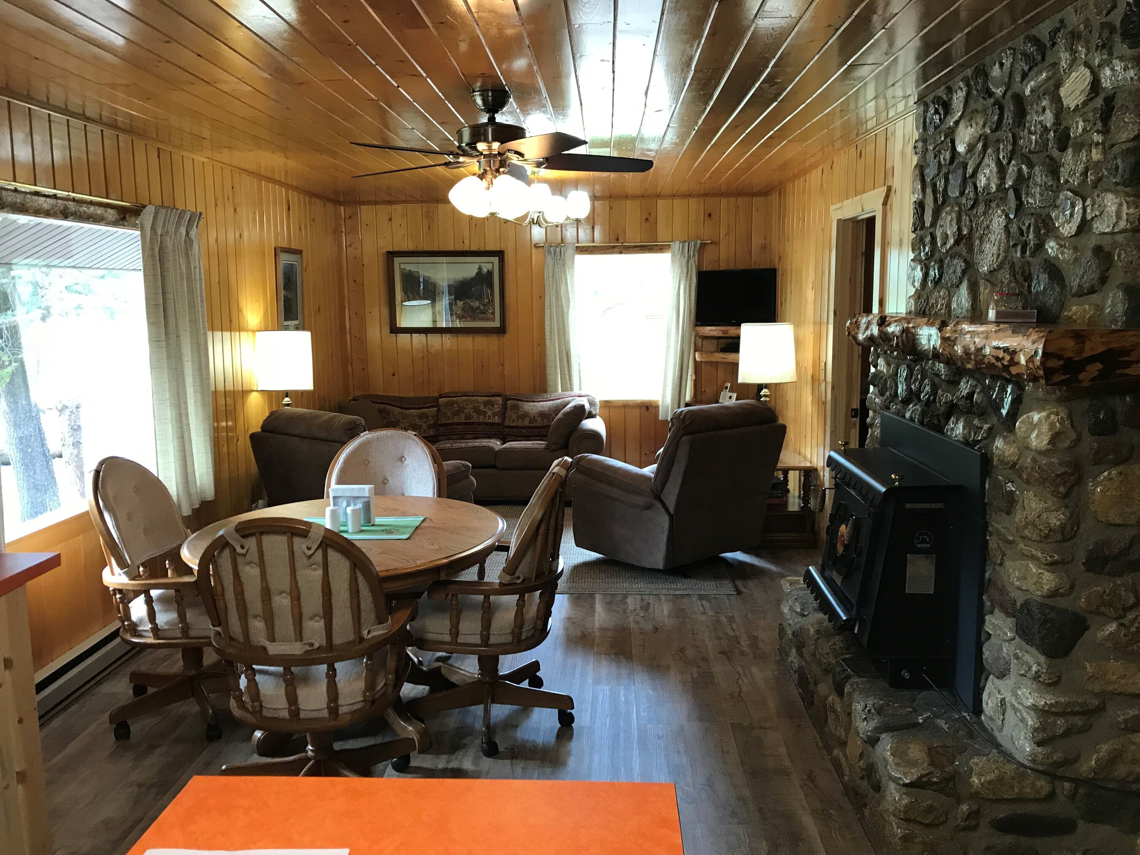 L’intérieur chaleureux d’un chalet avec une cheminée en pierre, des murs en bois, un ventilateur de plafond et un coin repas comprenant une table et des chaises.