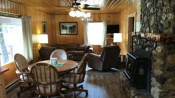 A cozy cabin interior with a stone fireplace, wooden walls, a ceiling fan, and a dining area with a table and chairs.