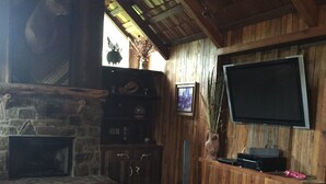 Ein rustikaler Innenraum mit Kamin aus Stein, Ledersofa, Holzwänden und einem angebrachten Fernseher.