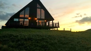 Una casa grande y moderna con un balcón destacado y ventanas amplias, ubicada en una colina cubierta de césped con un atardecer al fondo.