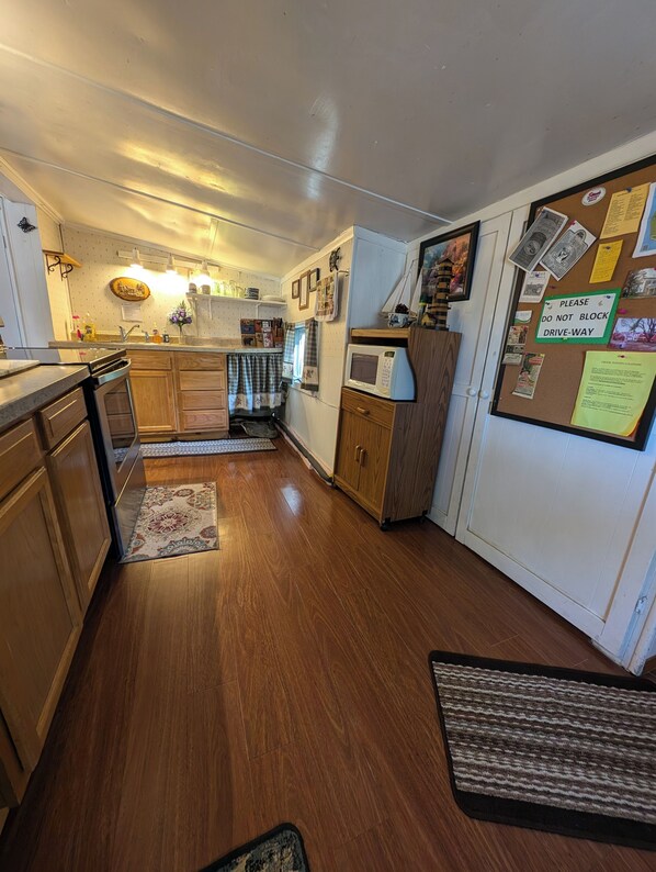 A compact kitchen with wooden cabinets, a microwave, and a corkboard with various notes and pictures.