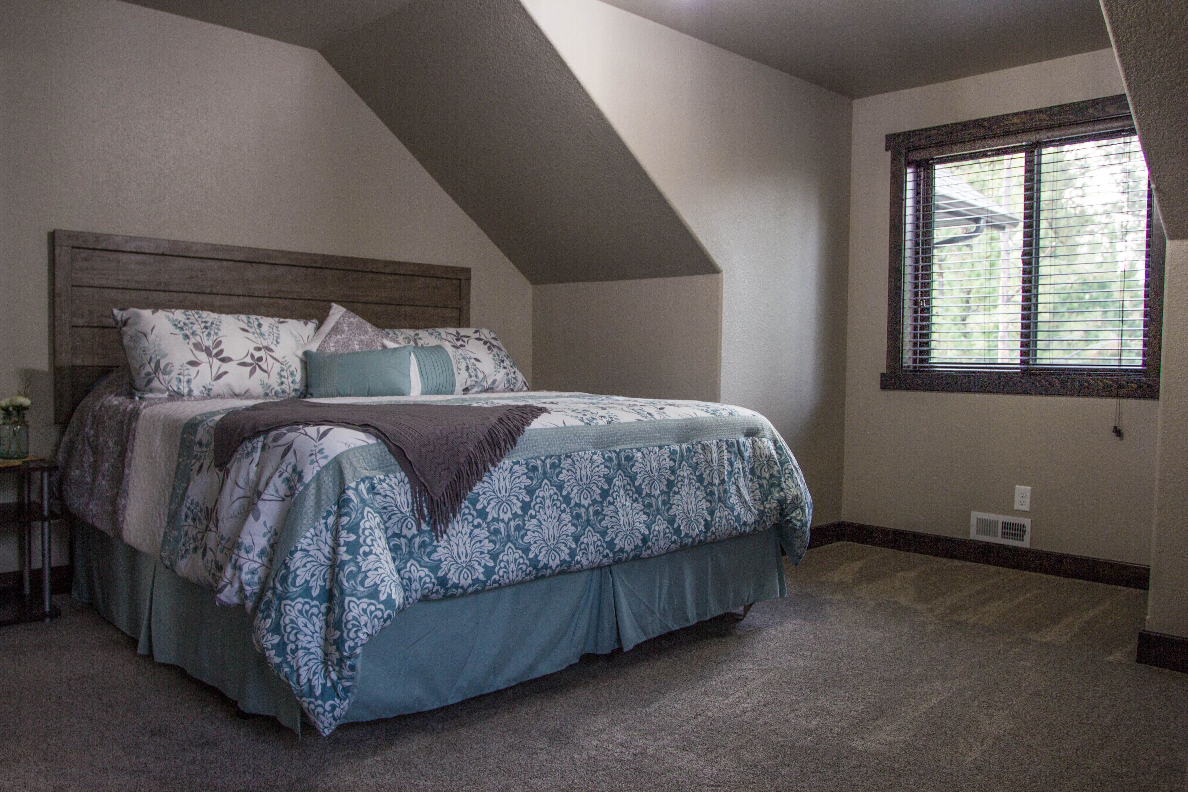A bedroom with a sloped ceiling, a large bed with patterned bedding, a nightstand, and a window with blinds.