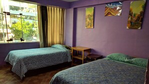 Una habitación con dos camas, una ventana con cortinas, una pequeña mesa de madera y una pared con tres cuadros.