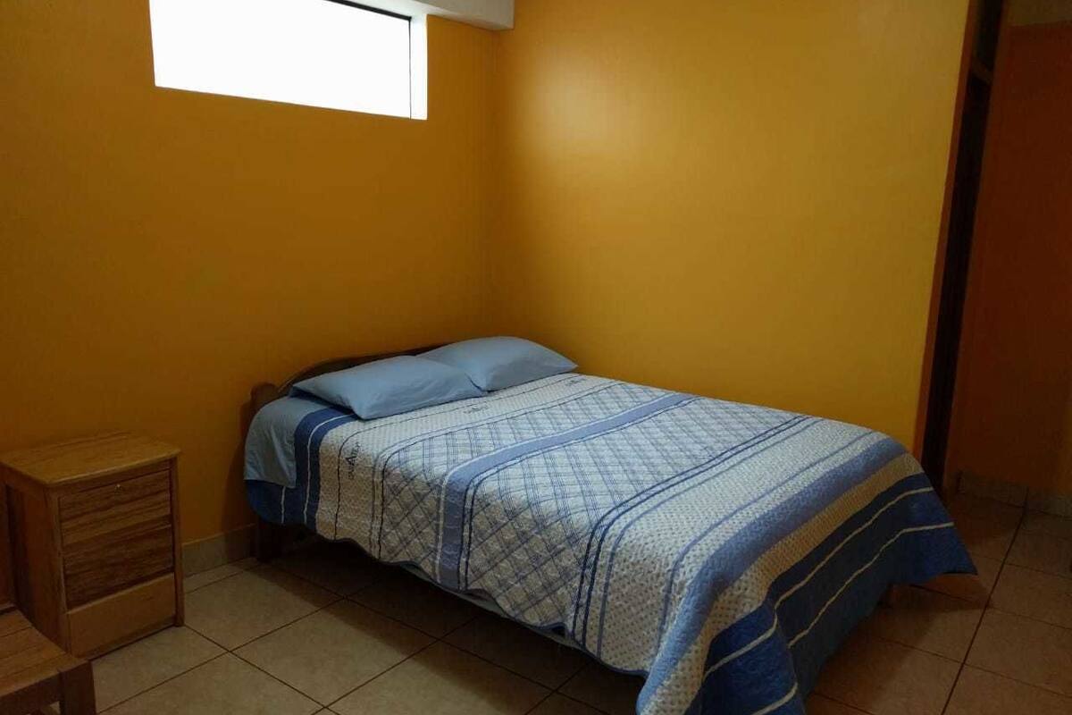 Une petite chambre avec un lit simple, une couvre-lit à rayures bleues et blanches, une table de chevet en bois et une fenêtre avec un store blanc.