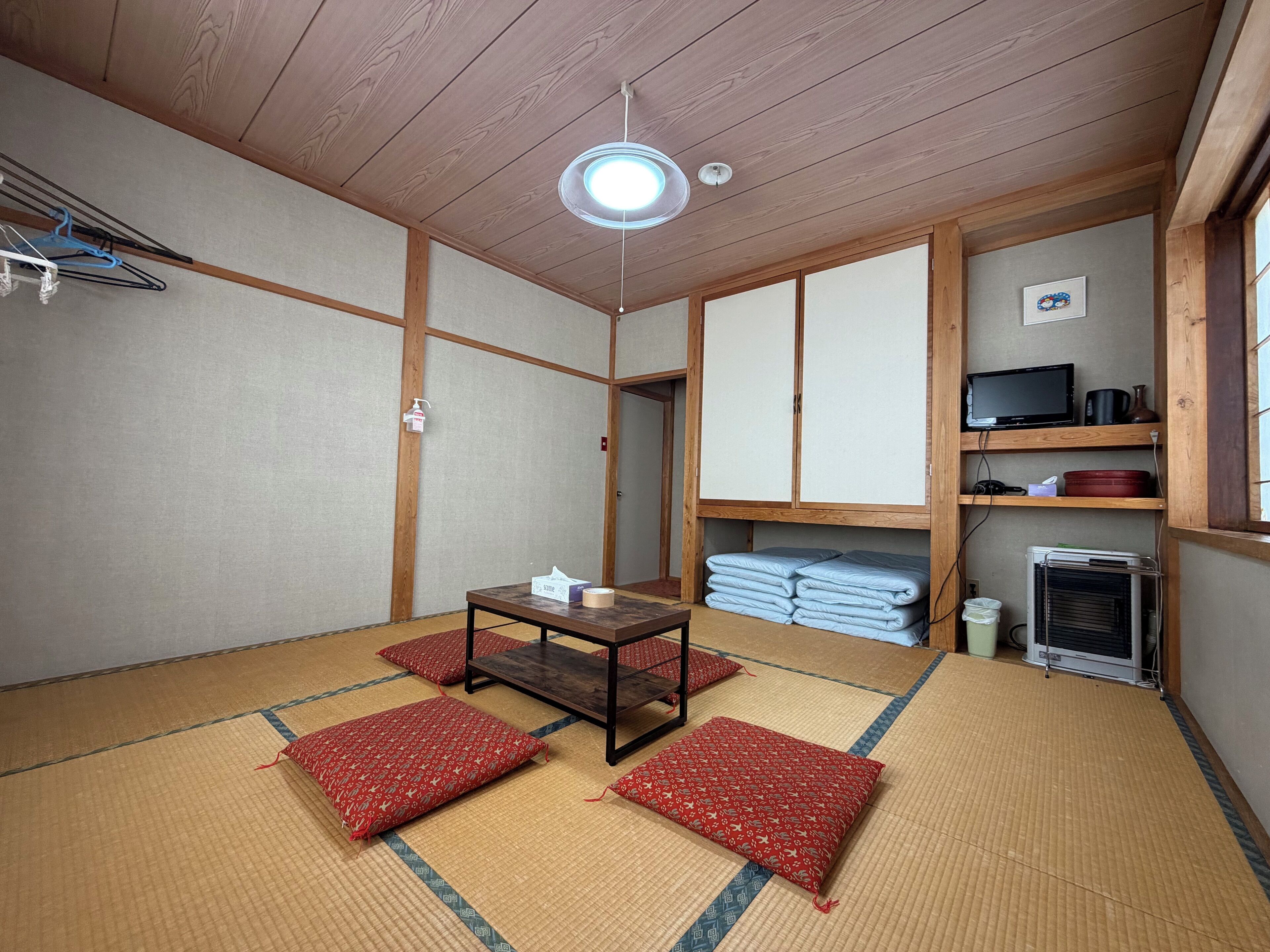 A traditional Japanese room with tatami flooring, a low table, cushions, a television, and a heater.