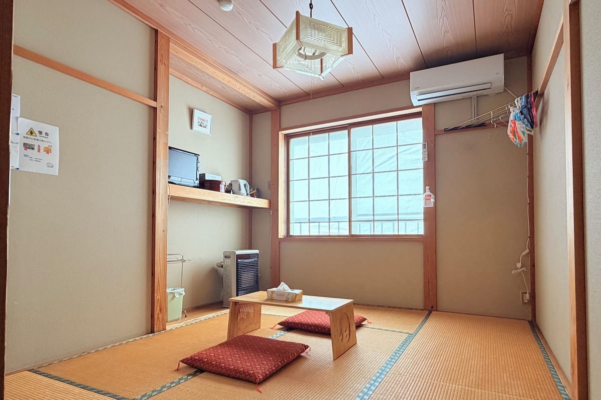 Ein traditioneller japanischer Raum mit Tatami-Boden, einem Futonbett, einem Schreibtisch mit Computer und einem großen Fenster.