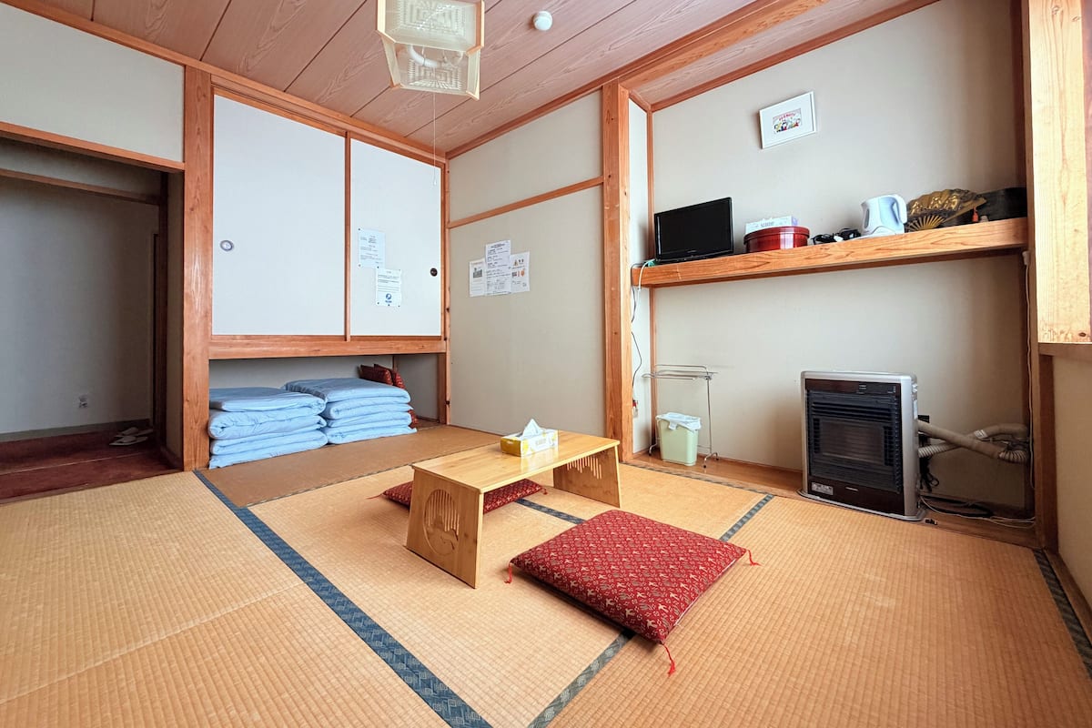 Ein traditioneller japanischer Raum mit Tatami-Boden, einem niedrigen Holztisch, einem roten Kissen, einem Fernseher und einer Heizung.