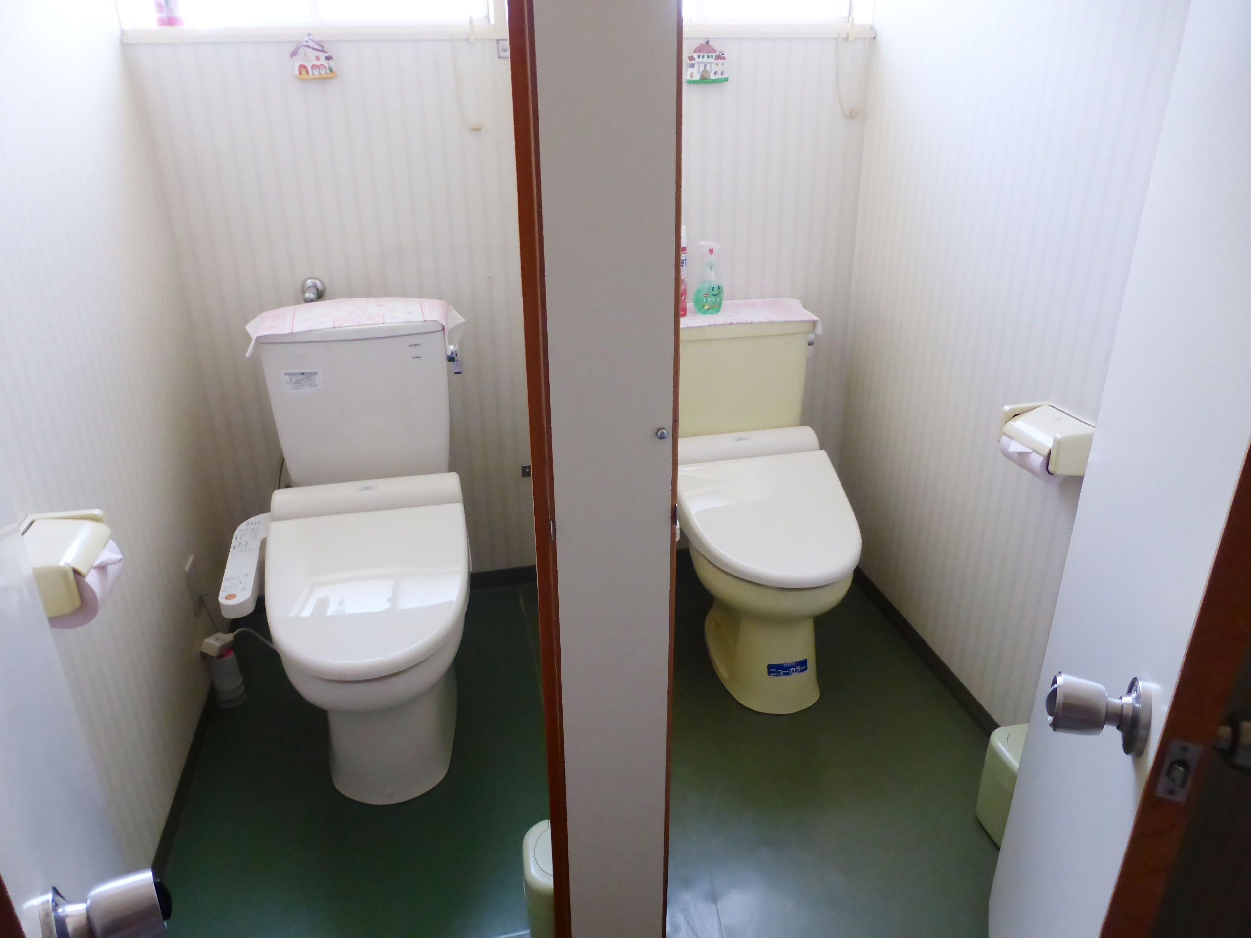 A small, narrow bathroom with two toilets, a hand dryer, and a toilet paper holder.