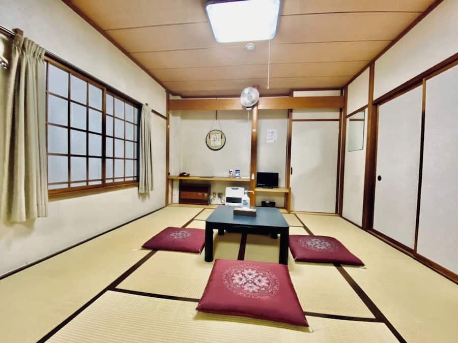 Una habitación japonesa tradicional con suelo de tatami, una mesa baja, cojines, un televisor y un ventilador de techo.
