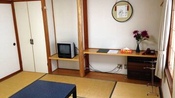 Una habitación tradicional japonesa con suelo de tatami, una mesa baja de tatami, un televisor en un estante, un pequeño escritorio con un jarrón de flores y una obra de arte colgada.