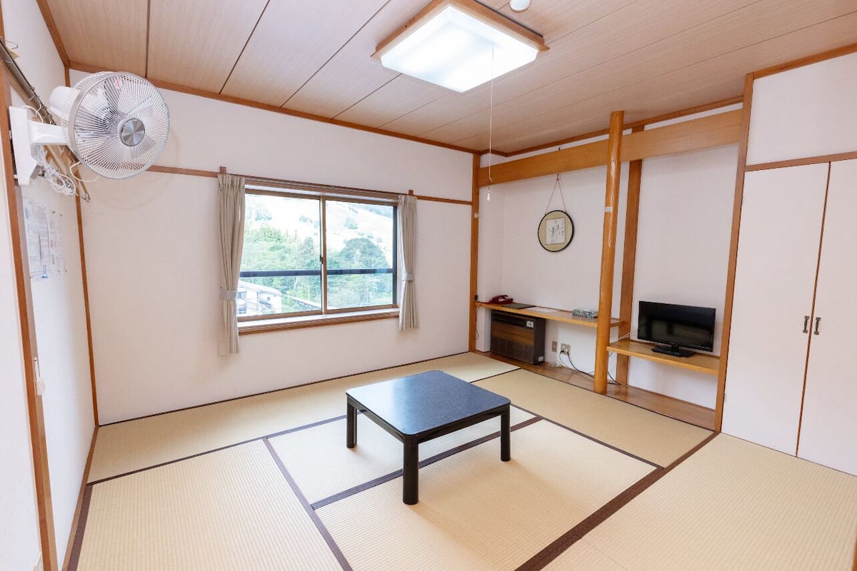 A traditional Japanese room with tatami flooring, a tatami mat table, a television on a wooden shelf, a fan, and a window with a view of greenery.