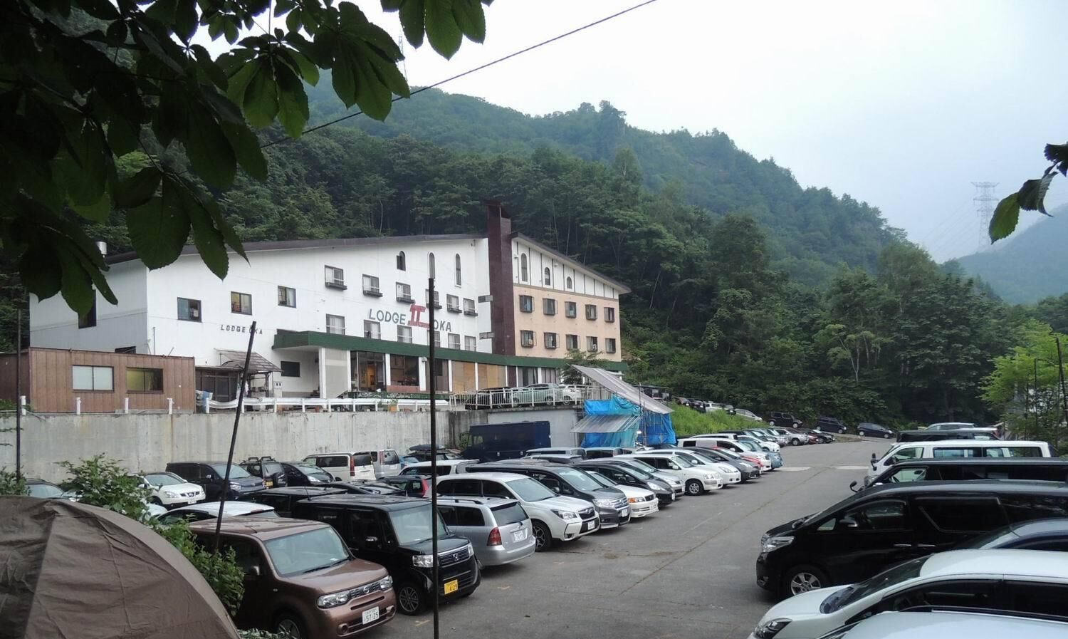 A parking lot with several cars, a building labeled 'Lodge Okawa', and a mountainous backdrop.