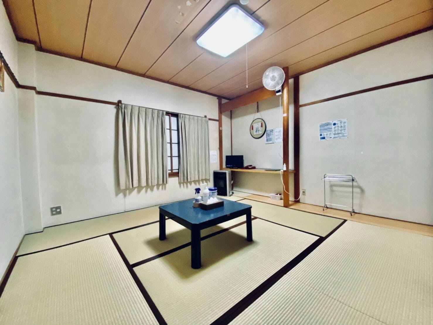 Una habitación japonesa tradicional con suelo de tatami, una mesa baja, un televisor en un estante y un abanico en la pared.