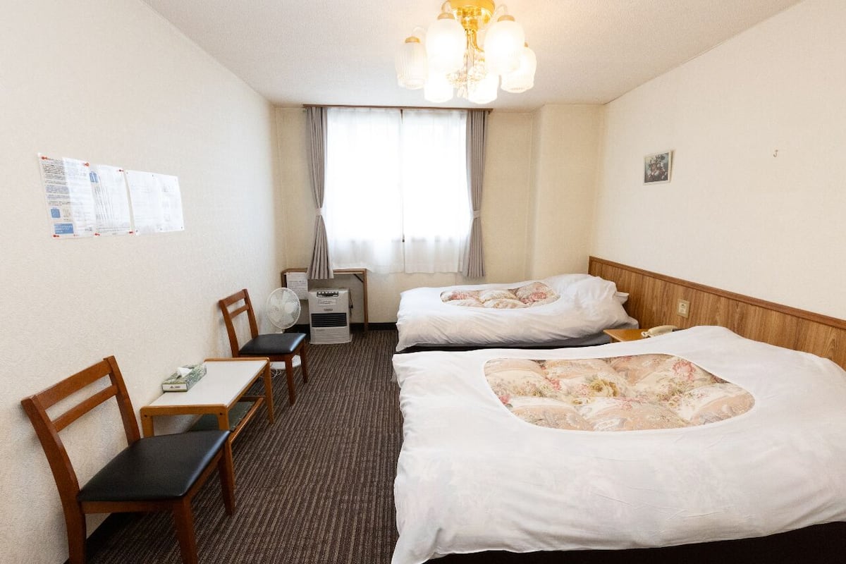 A hotel room with two beds, a desk, chairs, a fan, and a window with curtains.