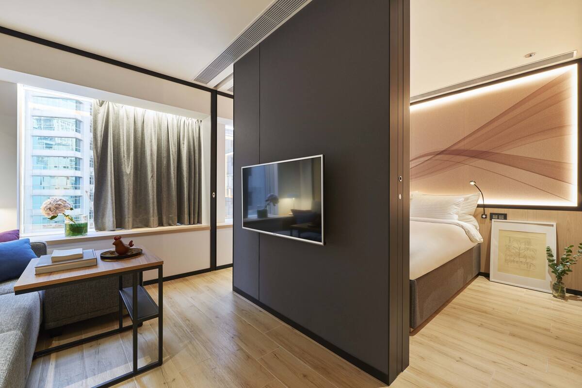 Moderni hotellihuone, jossa on suuri ikkuna, seinälle asennettu taulutelevisio, päätyosalla varustettu sänky sekä pieni pöytä, jossa on maljakko ja kirjoja.