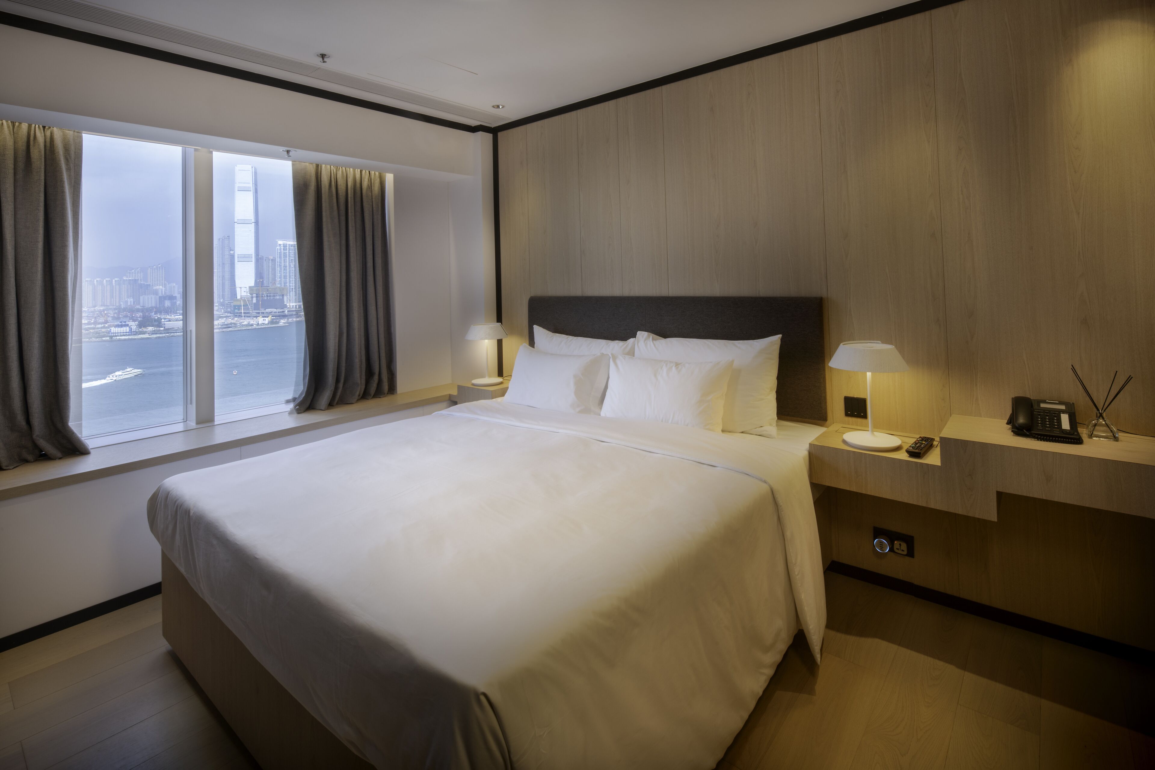 Una habitación de hotel con una cama grande, una mesita de noche con una lámpara, un teléfono y vistas a la ciudad a través de amplias ventanas.