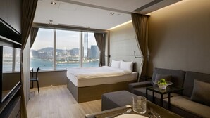 Een moderne hotelkamer met een groot bed, een sofa, een klein tafeltje met een schaal fruit, en uitzicht op een stadsbeeld en water.