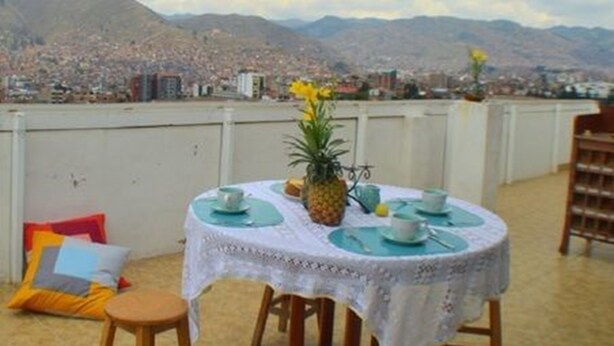 Un tavolo rotondo con tovaglia di pizzo bianco, apparecchiato con piatti, tazze e un centrotavola con ananas, con vista su una città e montagne.