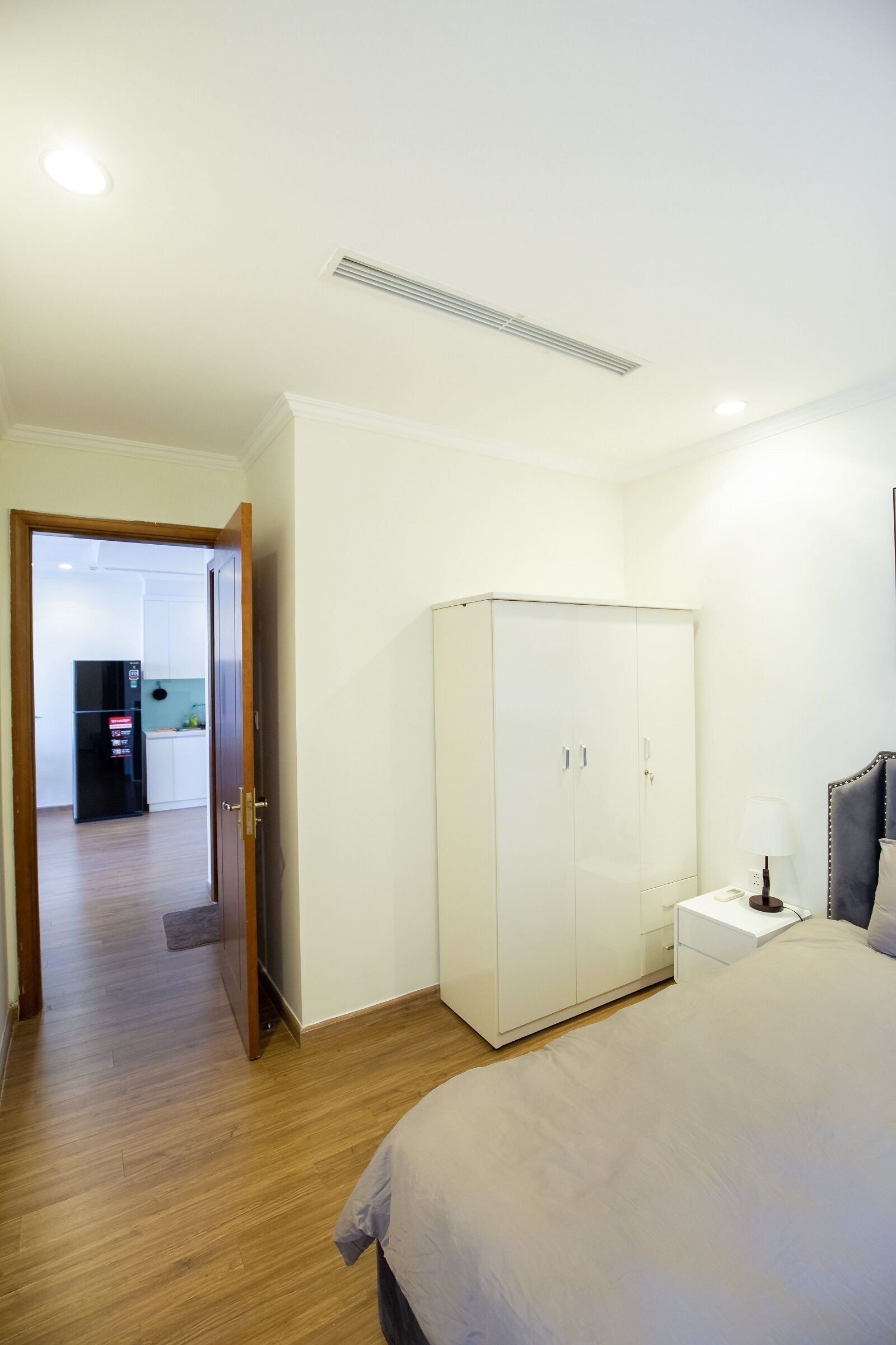 A bedroom with a bed, a wardrobe, a bedside table with a lamp, and a door leading to a kitchen area.