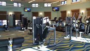 Un gimnasio con diversas máquinas de ejercicio, ventanas amplias y dos televisores en las paredes.