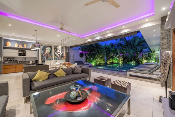 A modern living room with a glass coffee table, grey sofa, and a view of a pool and garden at dusk. - Villa Sawah (Seminyak)