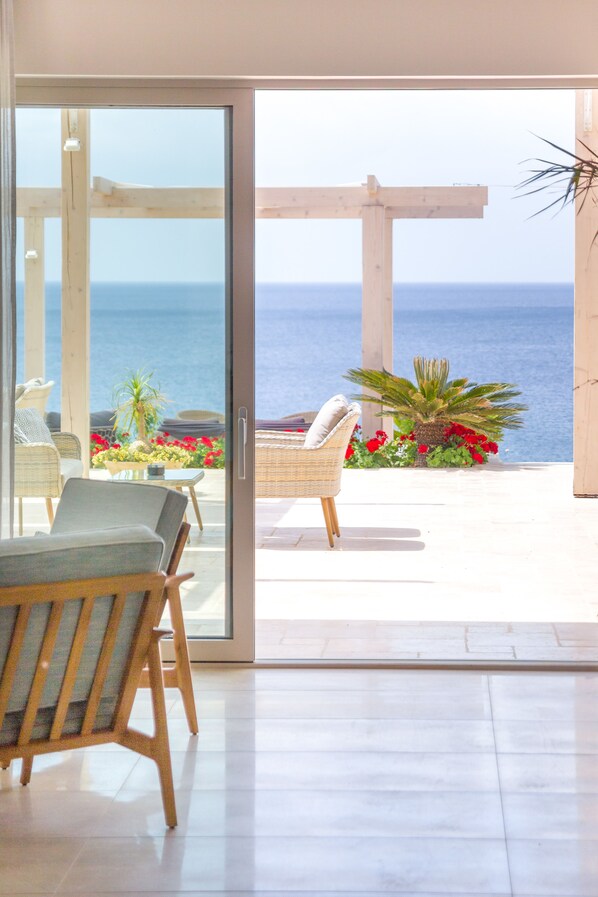 Habitación moderna con un amplio ventanal corredero que da a un balcón con vistas al mar. En el balcón, hay sillones de mimbre y una mesa pequeña con flores.