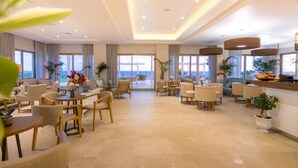 A spacious hotel lobby with multiple seating areas, large windows offering views of the outdoors, and a modern ceiling design. - King Maron Beach Hotel & Spa (Maroneia-Sapes)