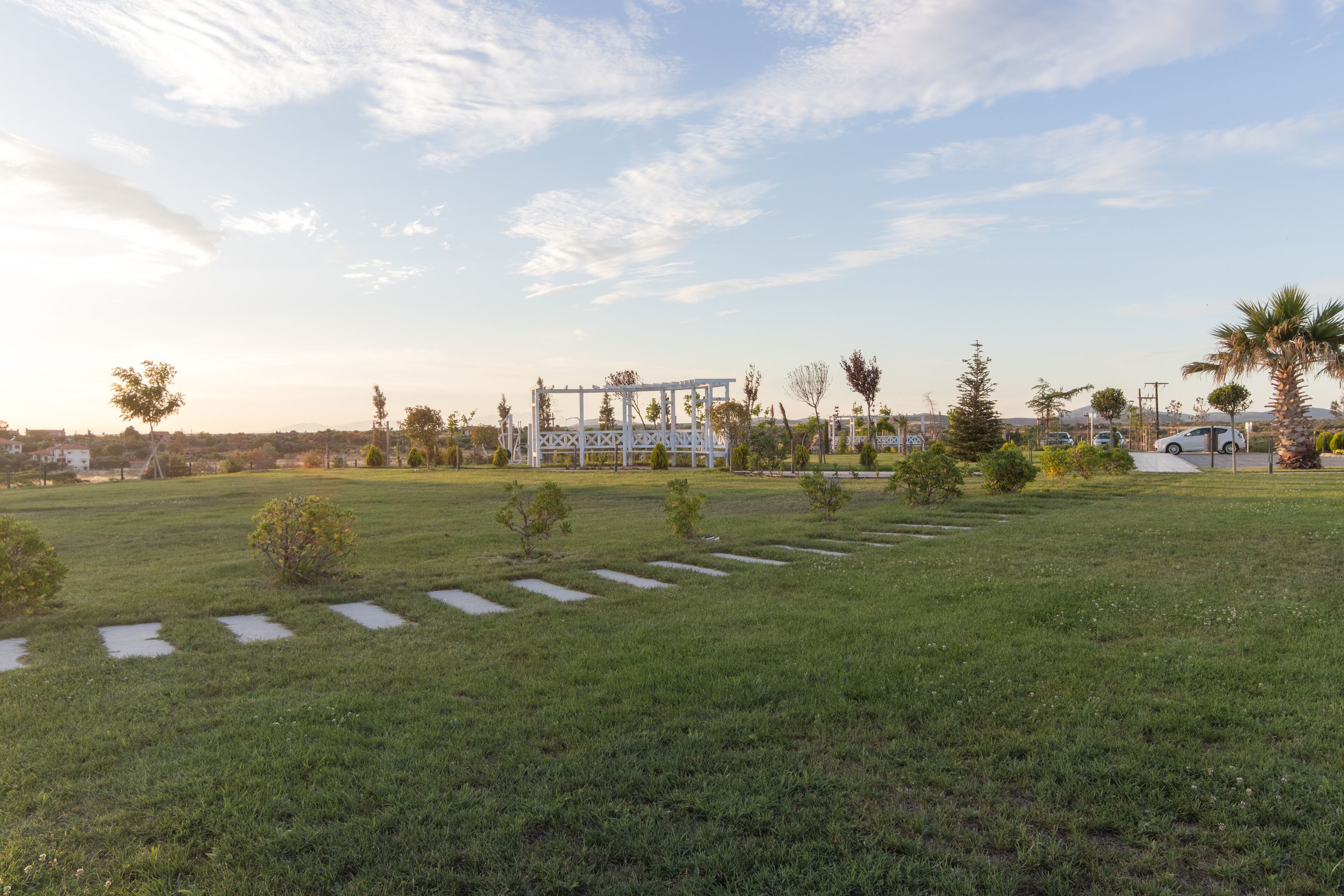 A grassy field with a white structure in the background, possibly a pavilion or gazebo, surrounded by trees and a few parked cars.