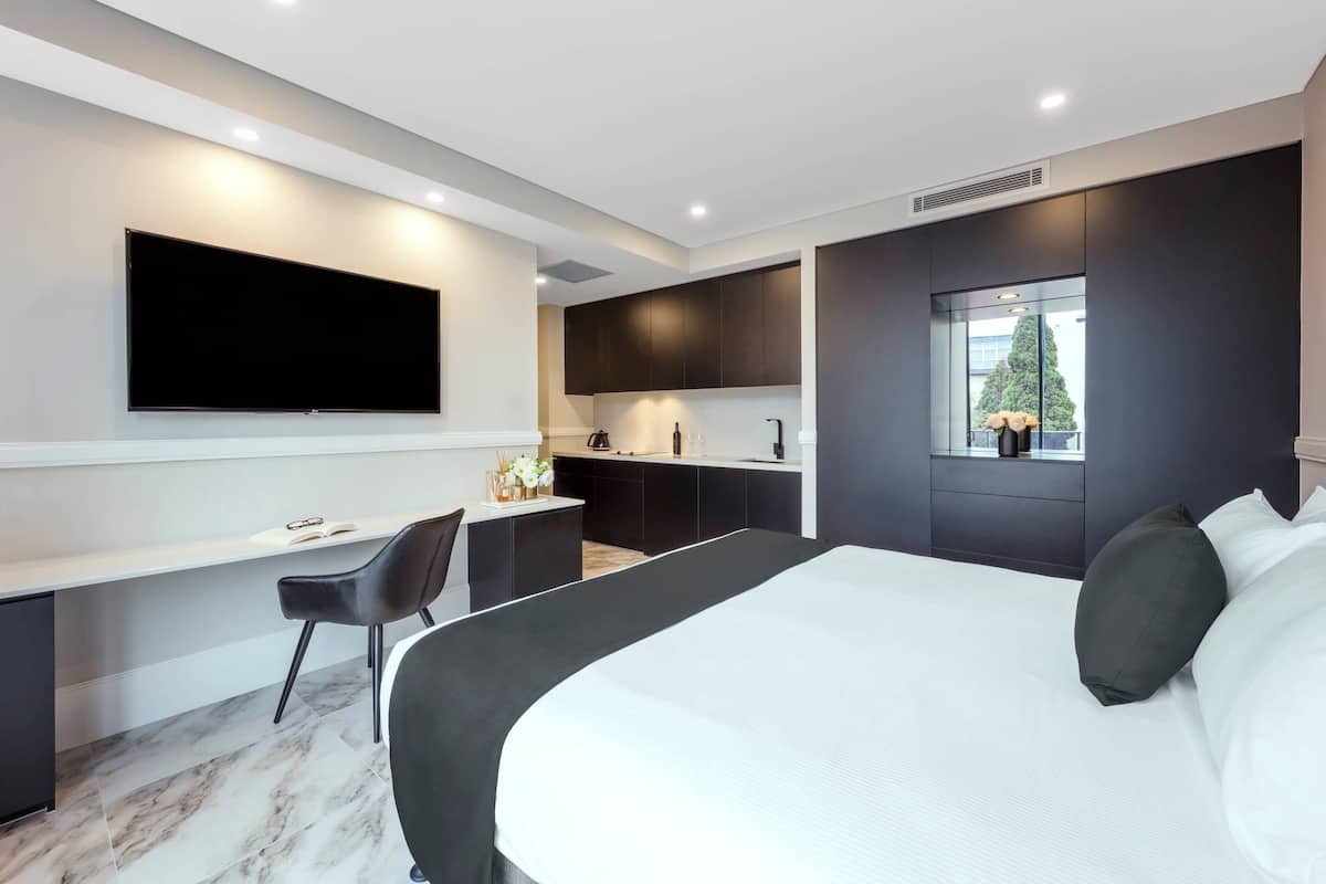 A modern hotel room with a large bed, a desk, a flat-screen TV, and a small kitchenette.