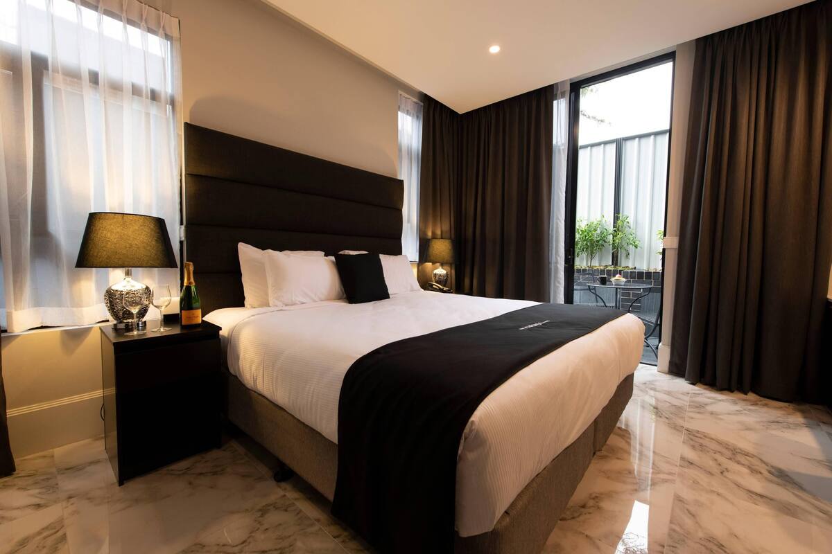 A modern hotel room with a large bed, dark headboard, and a view of the outdoor area.