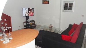 A living room with a TV, a glass coffee table, a black sofa with red cushions, and a red decorative panel. - Residence 2 Pièces Duplex (Abidjan)