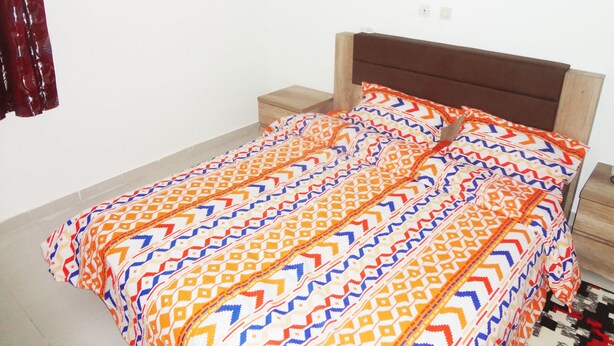 A bedroom with a bed, wooden headboard, nightstand, and a window with curtains. - Residence 2 Pièces Duplex (Abidjan)