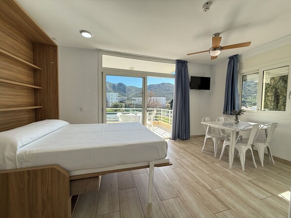 A bedroom with a bed, a dining table with chairs, a ceiling fan, and a view of the outdoors.