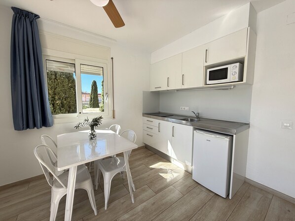 A modern kitchen with white cabinets, a microwave, a sink, and a small dishwasher. There is a dining table with chairs and a window with curtains.