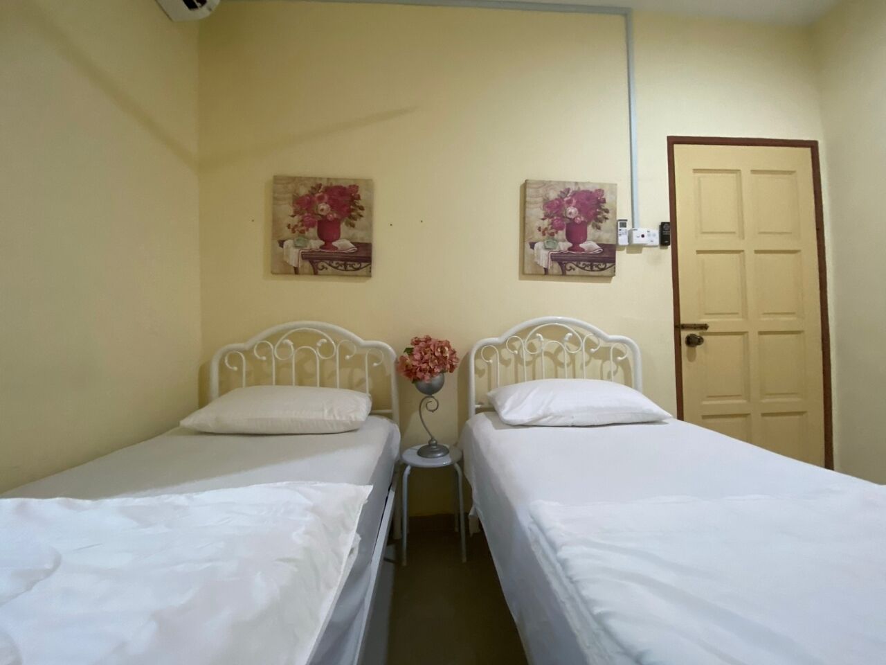 A room with two single beds, a small table with a vase, and a door.