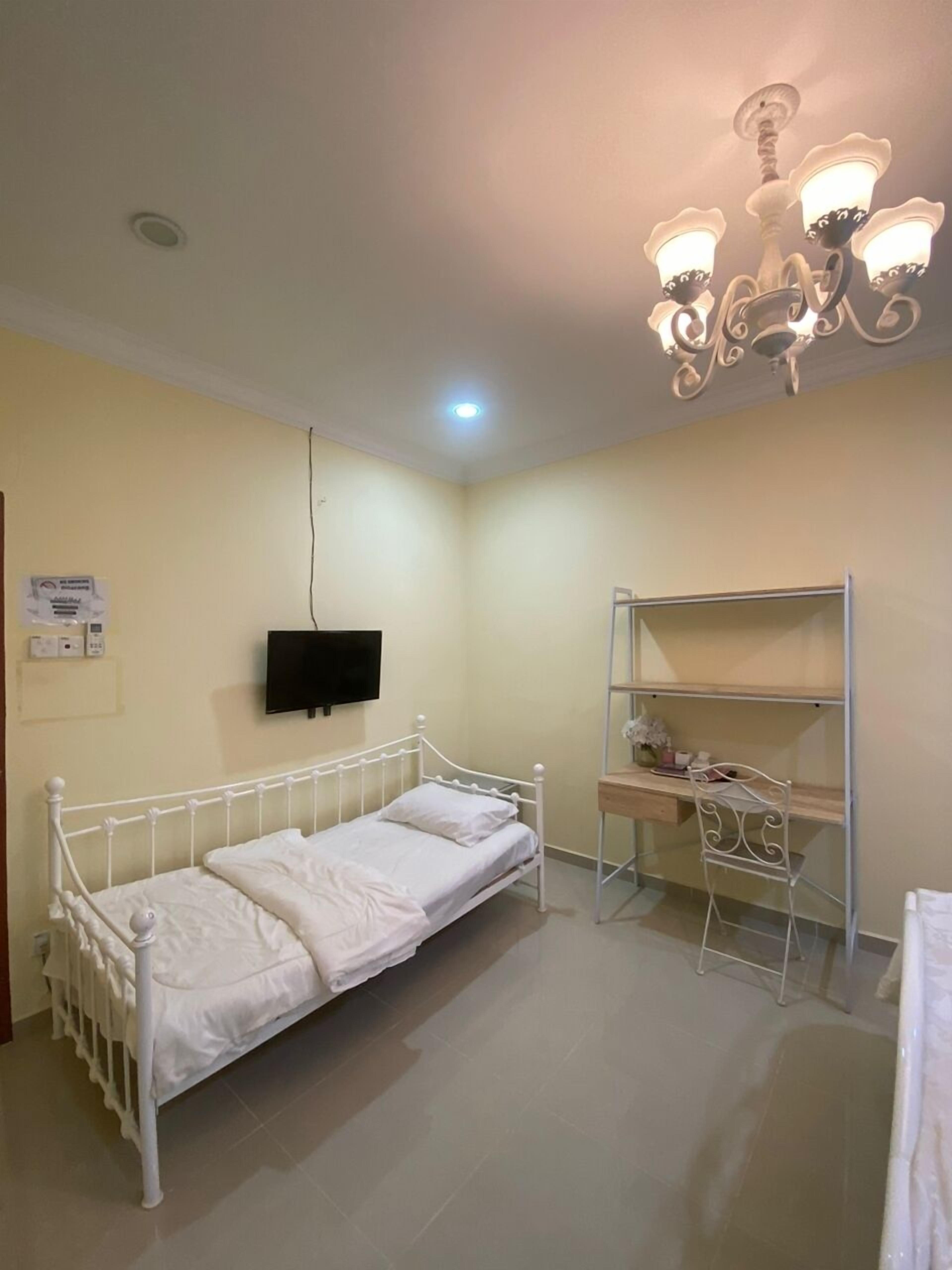 A small, well-lit room with a single bed, a desk with a chair, a TV mounted on the wall, and a decorative ceiling light fixture.