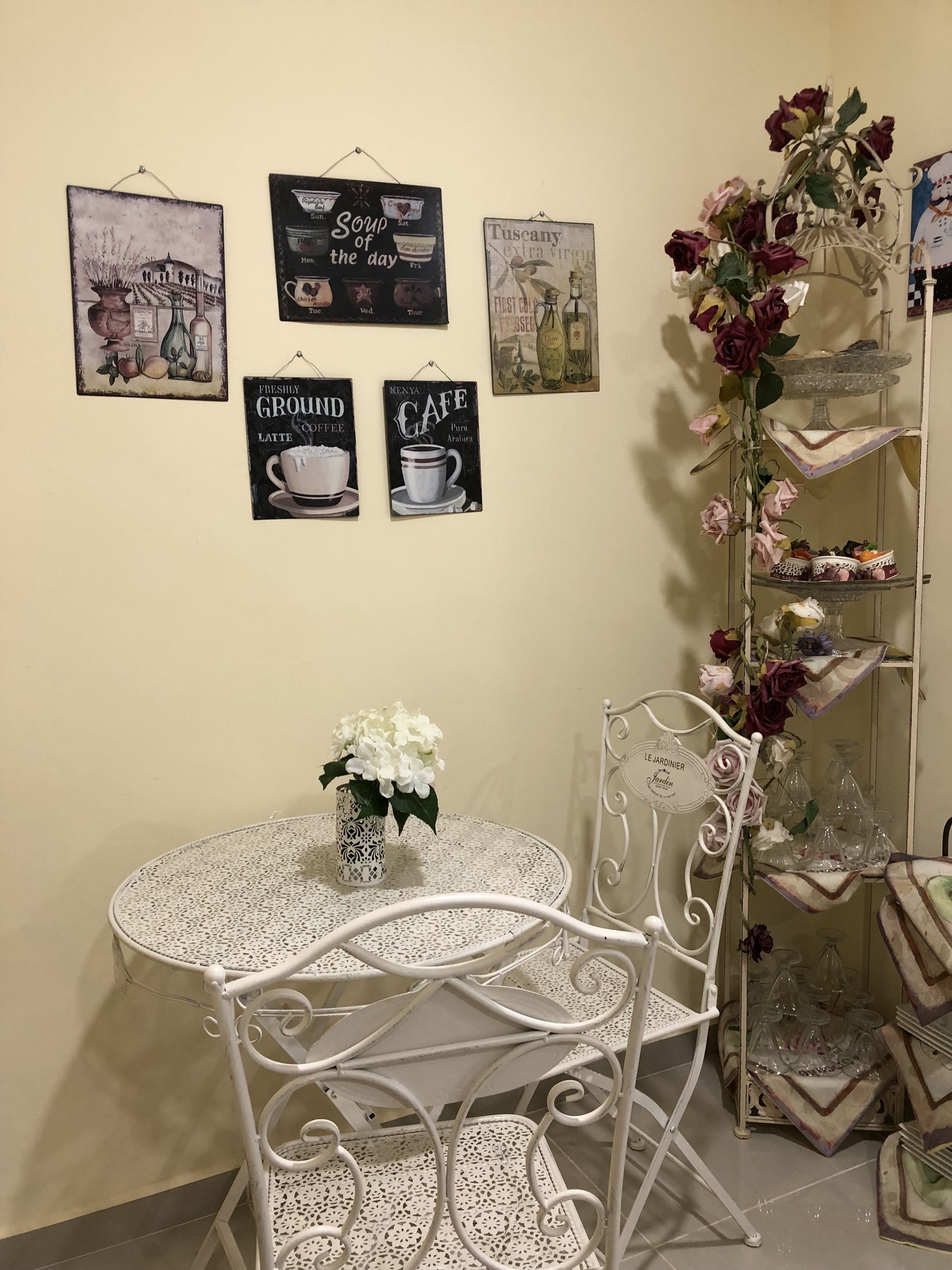 Een eethoek met een ronde tafel en stoelen, een vaas met witte bloemen, en wanddecoraties waaronder borden met 'Soep van de dag' en 'Kenya Cafe'.