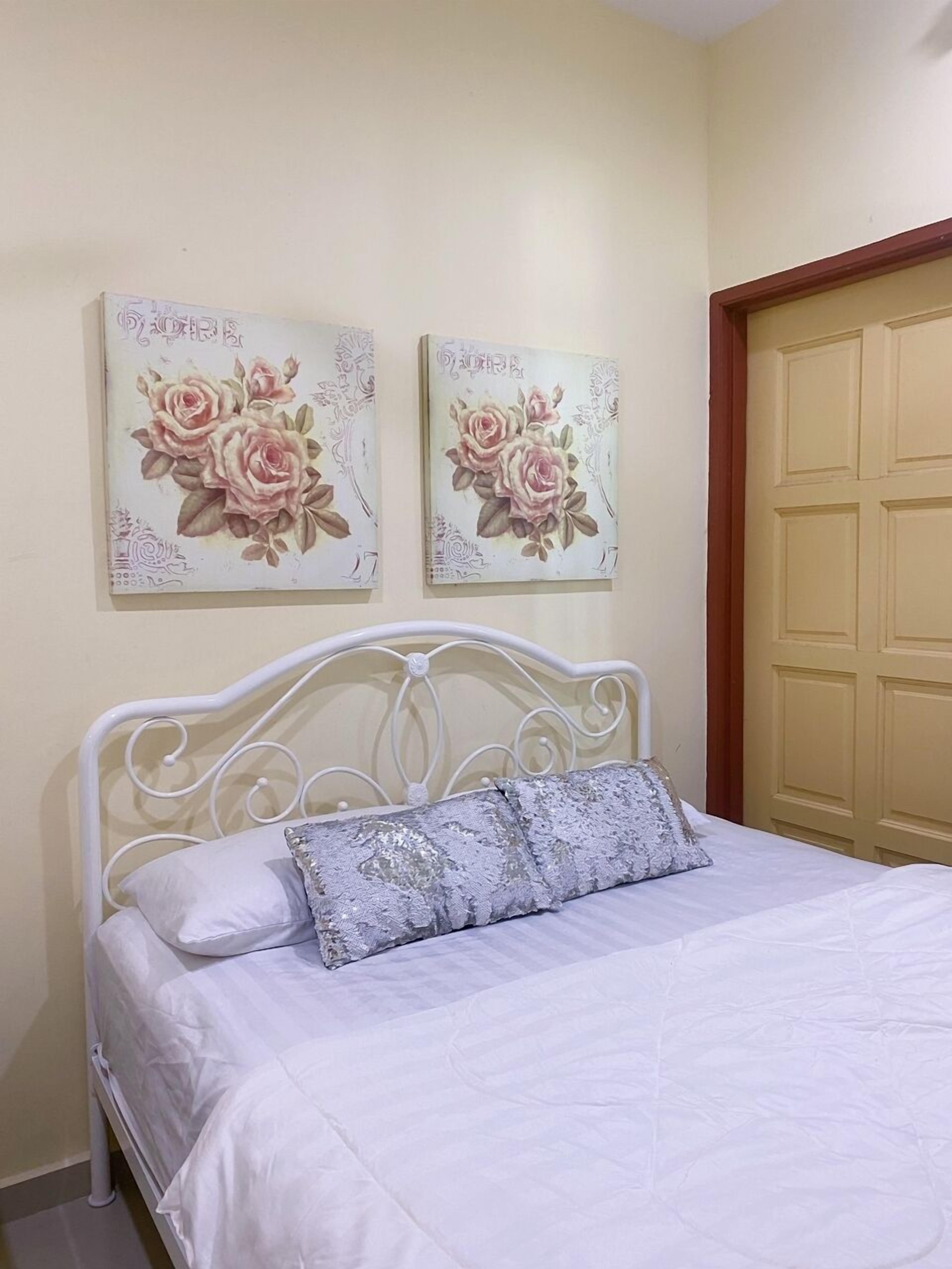 Een slaapkamer met een bed met een wit metalen frame, een witte spreidek en een decoratief kussen. Boven het bed hangen twee bloemrijke schilderijen aan de muur.