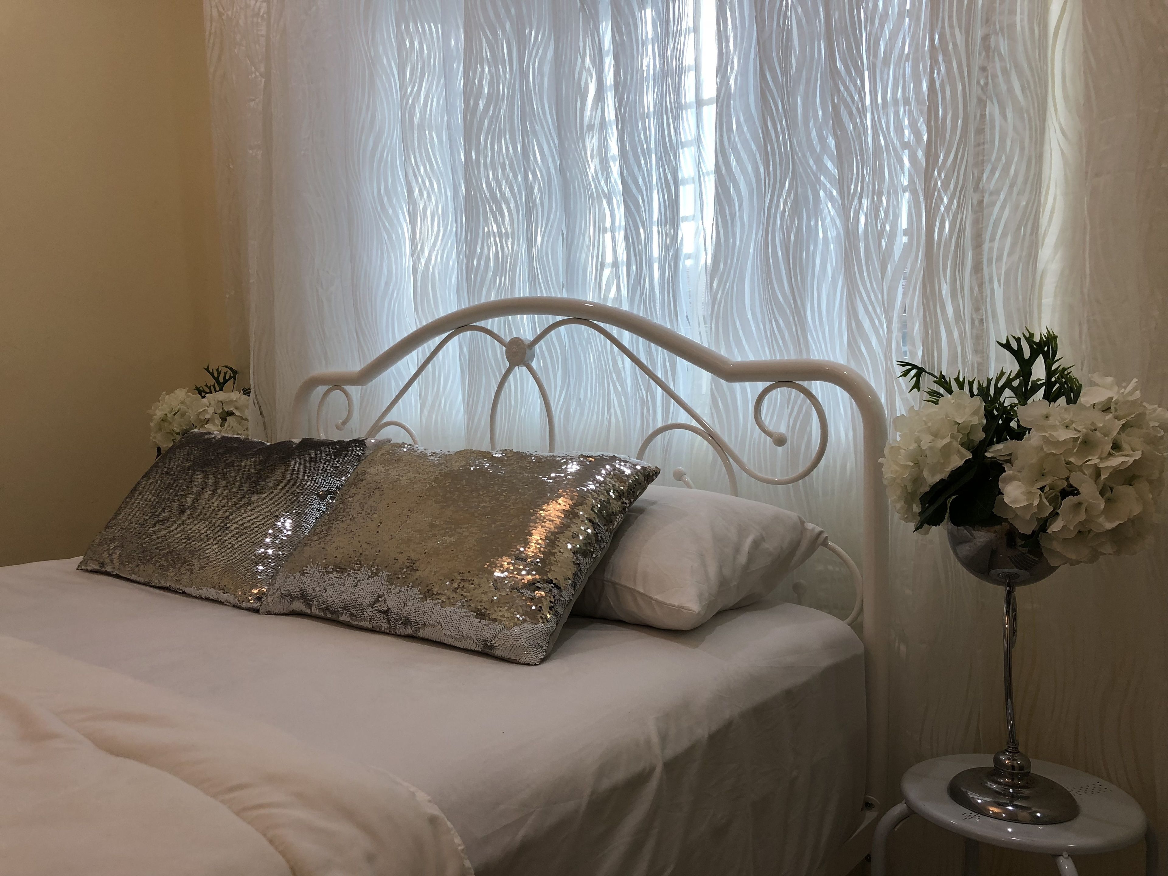 A bed with a metal frame, white bedding, and decorative pillows. There is a bedside table with a vase of flowers.