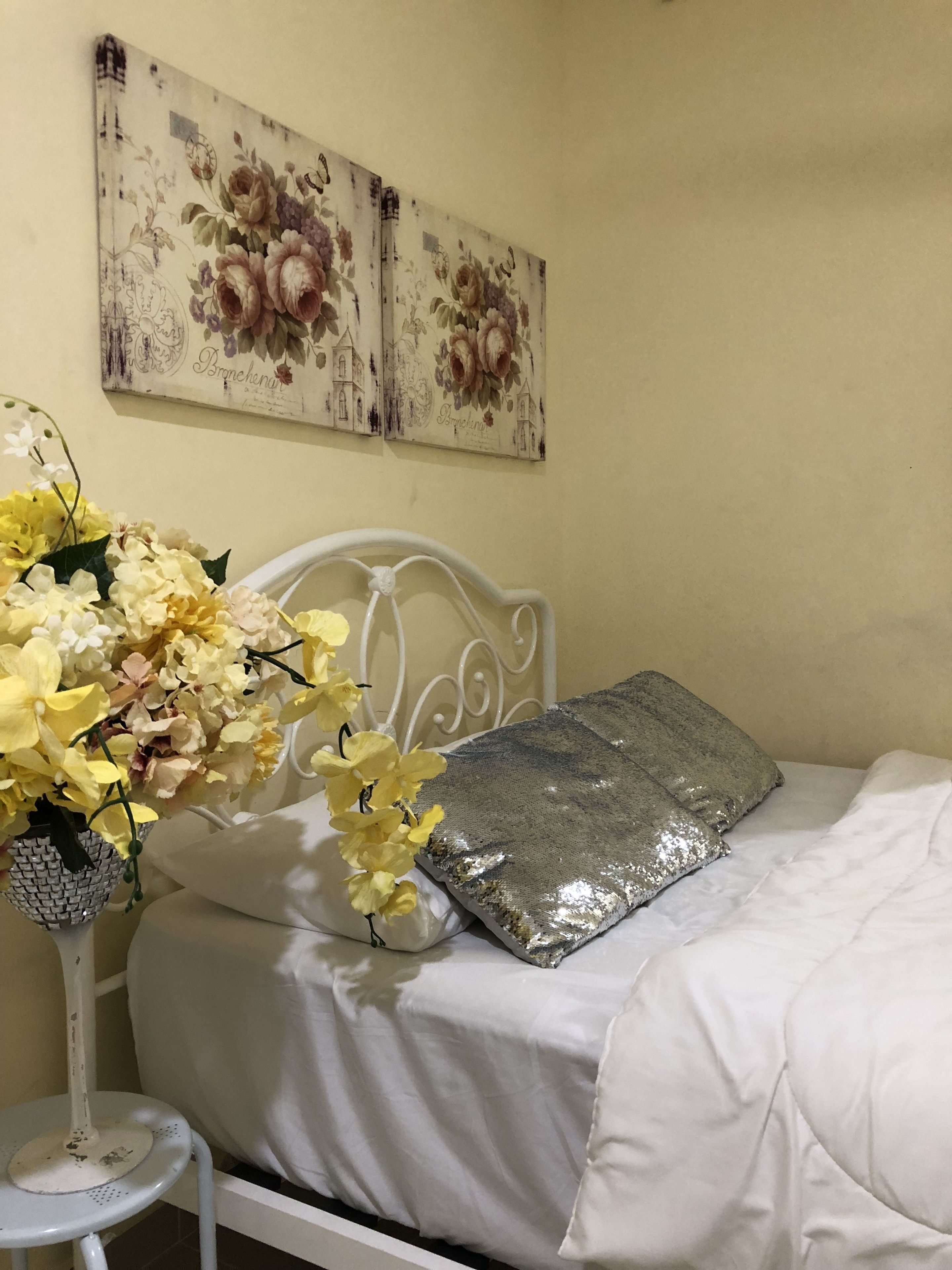 A bed with a white headboard, a silver pillow, and a vase of yellow flowers on a white table beside it.