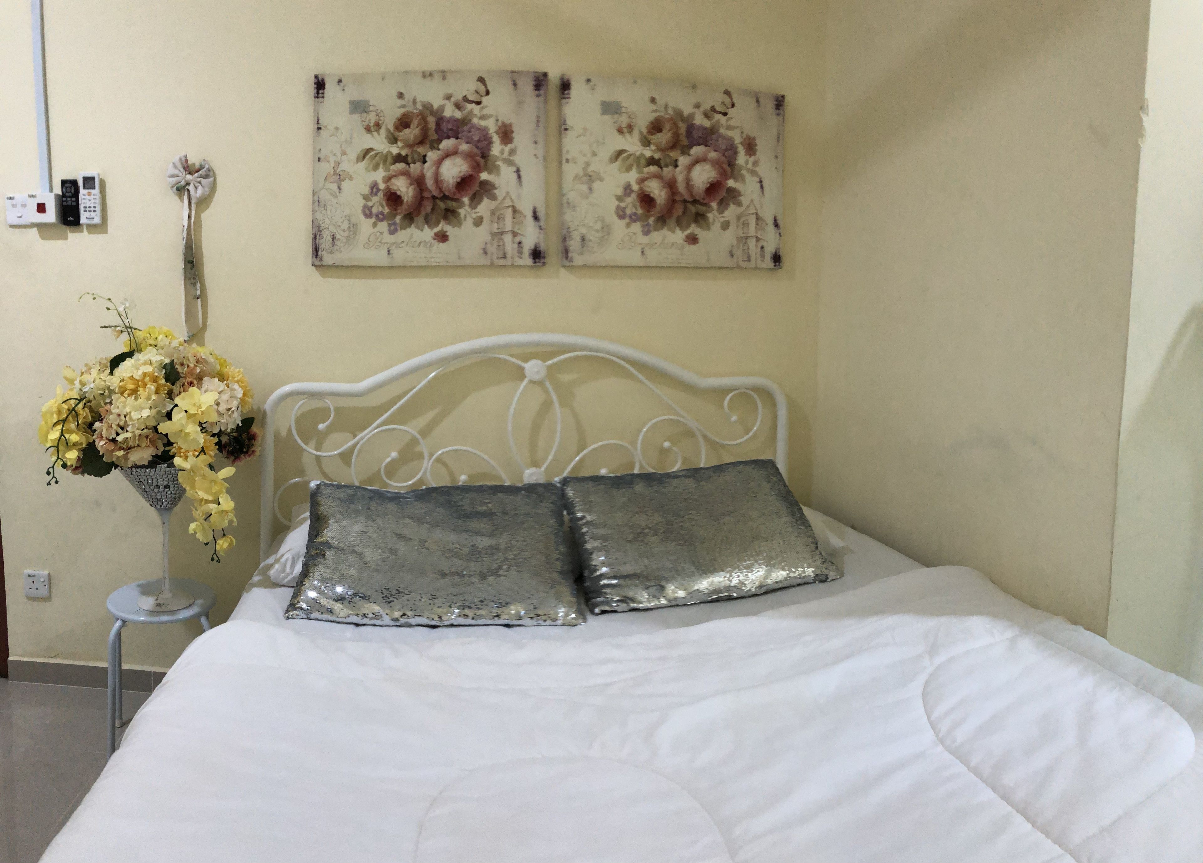 A bed with white bedding and two metallic pillows, a small white metal stand with a vase of flowers, and two floral wall art pieces above the bed.