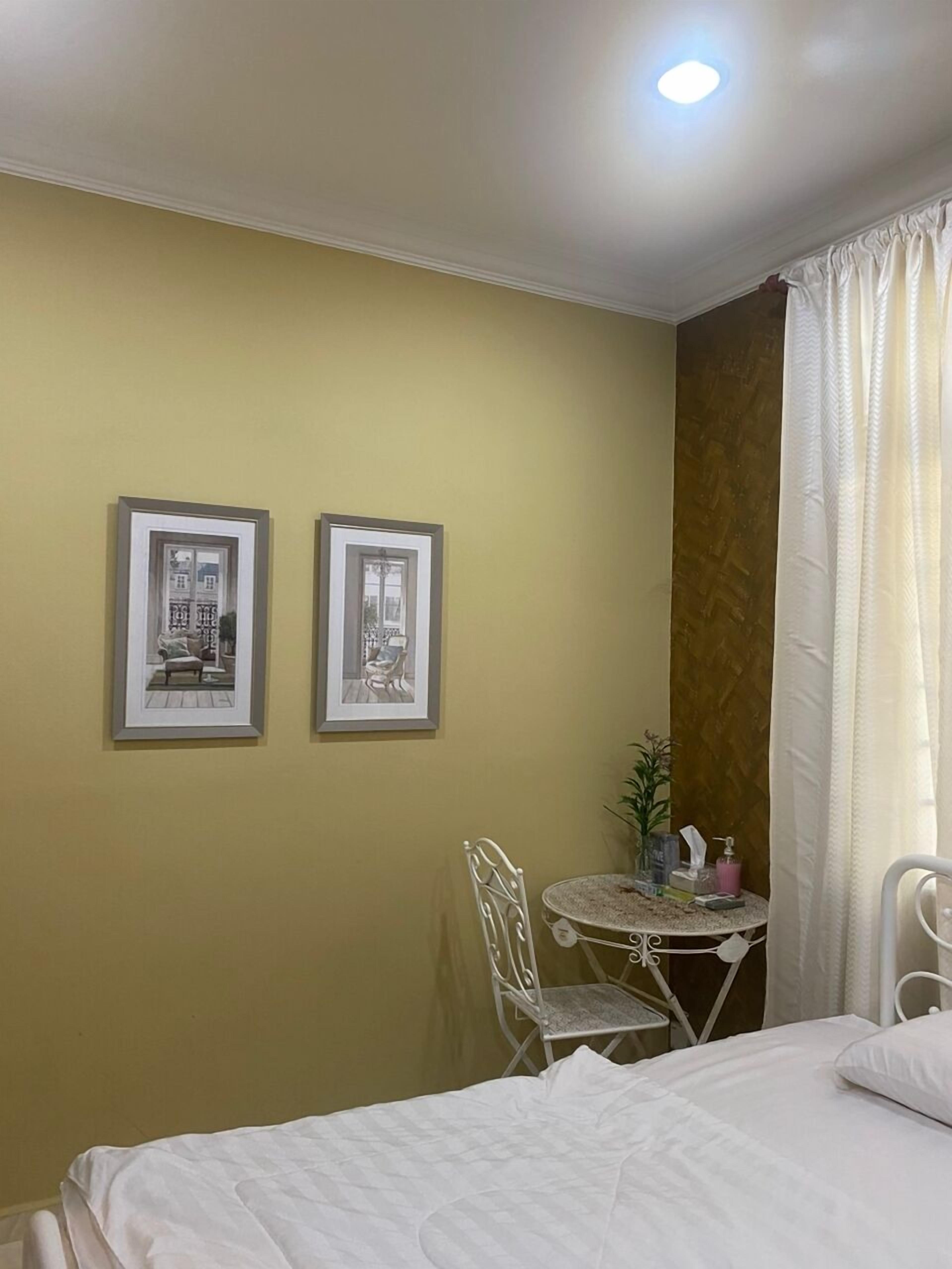 A hotel room with a bed, a small round table with a chair, two framed pictures on the wall, and a window with curtains.