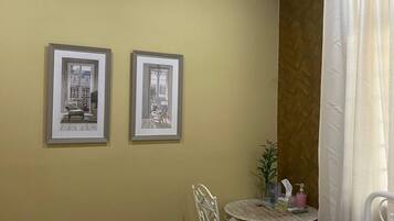 A hotel room with a bed, a small round table with a chair, two framed pictures on the wall, and a window with curtains.